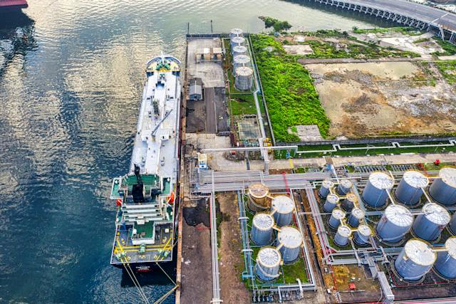 Aerial view of a large white tanker ship docked at a pier alongside cylindrical storage tanks within a petroleum facility, with lush greenery on one side and a reflective water body on the other.